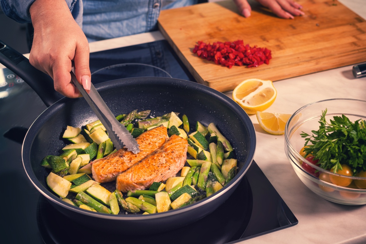 Eine Person brät in einer Pfanne Lachsfilets zusammen mit grünem Spargel und Zucchini an.
