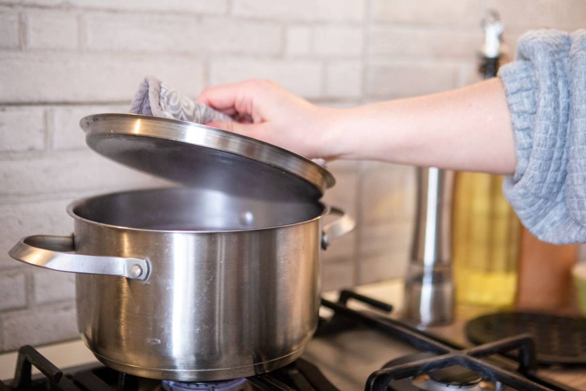 Person hebt den Deckel eines Edelstahltopfs auf einem Gasherd, während Gemüse gedünstet wird.
