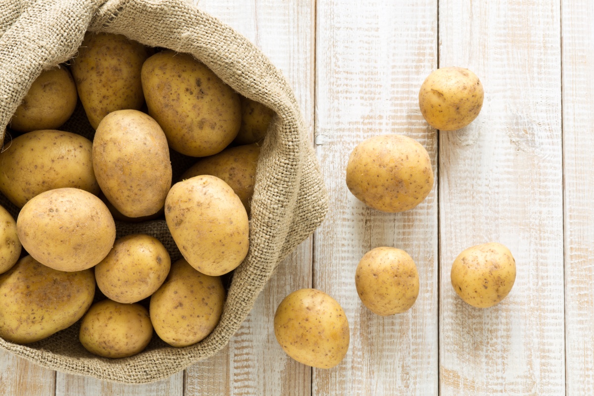 Ein offener Jutesack mit Kartoffeln auf einem hellen Holztisch, einige Kartoffeln liegen verstreut daneben.
