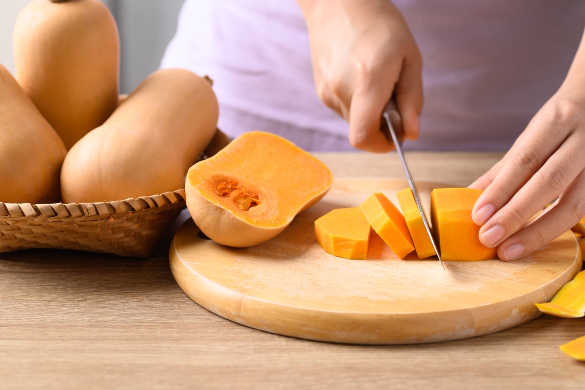 Person schneidet Butternut-Kürbis auf einem Holzbrett in Scheiben, daneben liegen ganze Kürbisse.