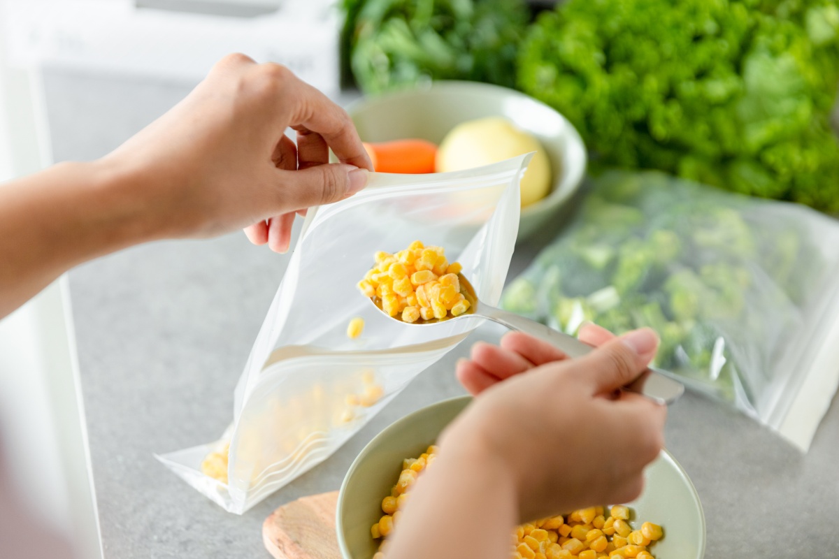 Person füllt Mais mit einem Löffel in einen Gefrierbeutel. Verschiedenes Gemüse im Hintergrund.