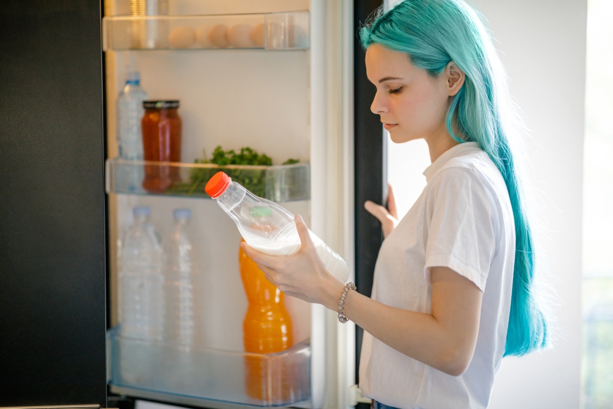 Frau mit türkisfarbenem Haar hält eine Milchflasche vor einem offenen Kühlschrank.