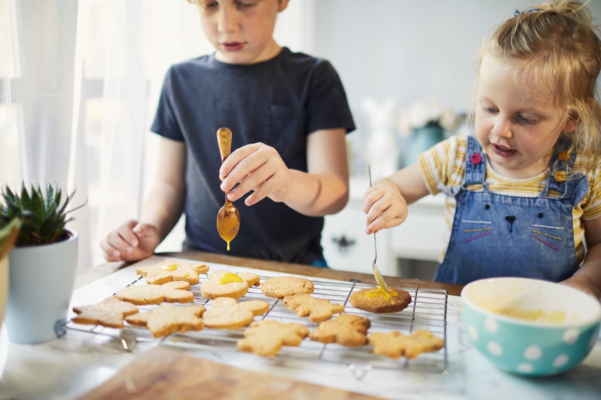 Zwei Kinder verzieren Plätzchen mit gelbem Zuckerguss, während die Plätzchen auf einem Abkühlgitter liegen.