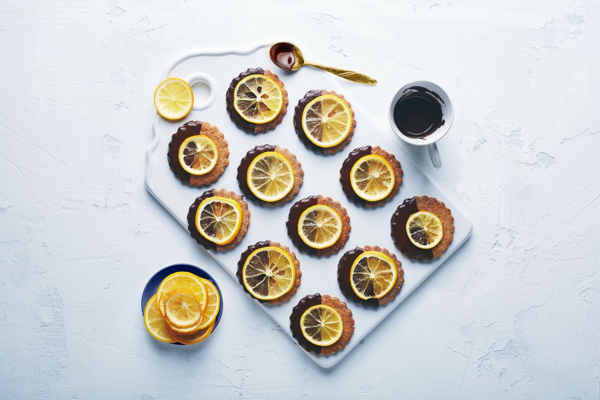 Frisch verzierte Zitronenplätzchen, daneben geschnittene Zitronenscheiben und eine Tasse mit Schokoglasur.