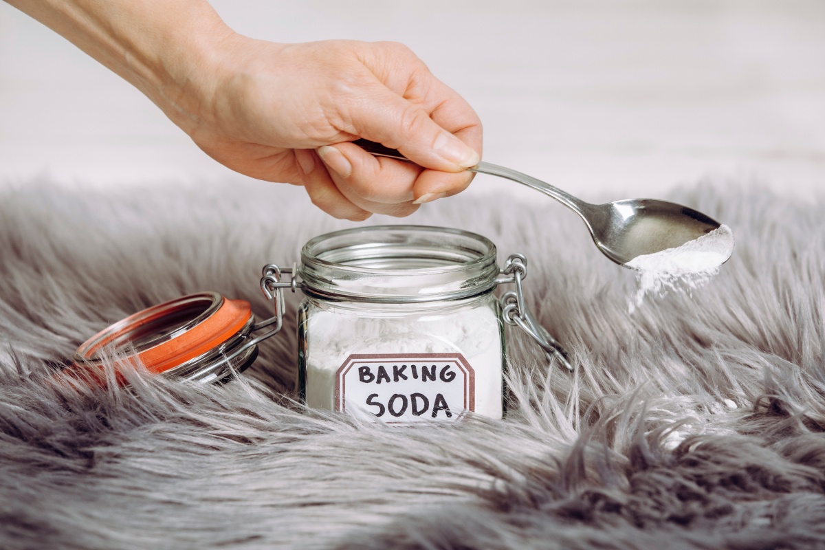 Eine Person streut mit einem Löffel Natron auf einen grau melierten Langhaar-Teppich.