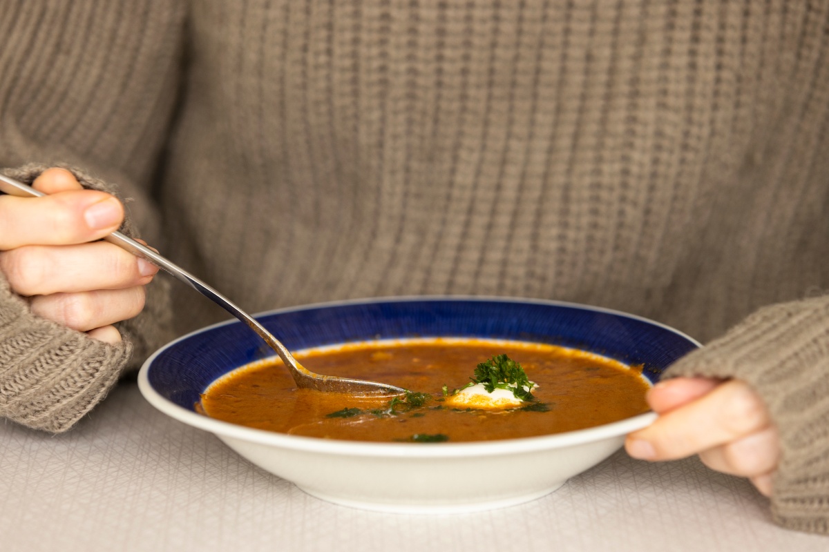 Person hält einen Löffel in einen Teller mit heißer Suppe, garniert mit Sahne und Kräutern.