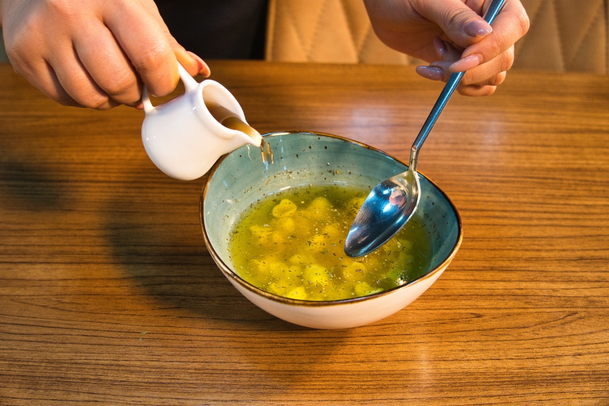 Person gießt Soße aus einer kleinen Kanne in eine Schüssel mit Suppe, hält dabei einen Löffel in der anderen Hand.
