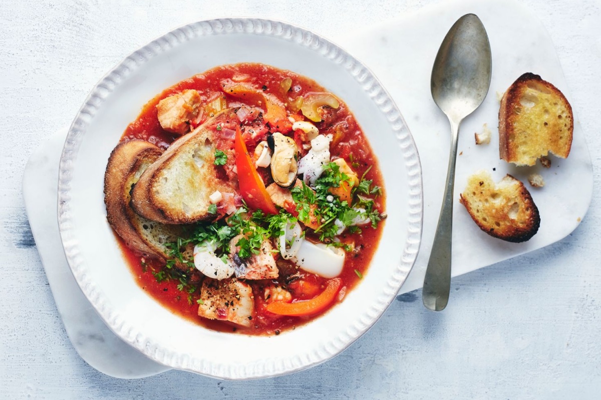 Weißer Teller mit einer tomatigen Fischsuppe, daneben ein Löffel und geröstetes Brot.