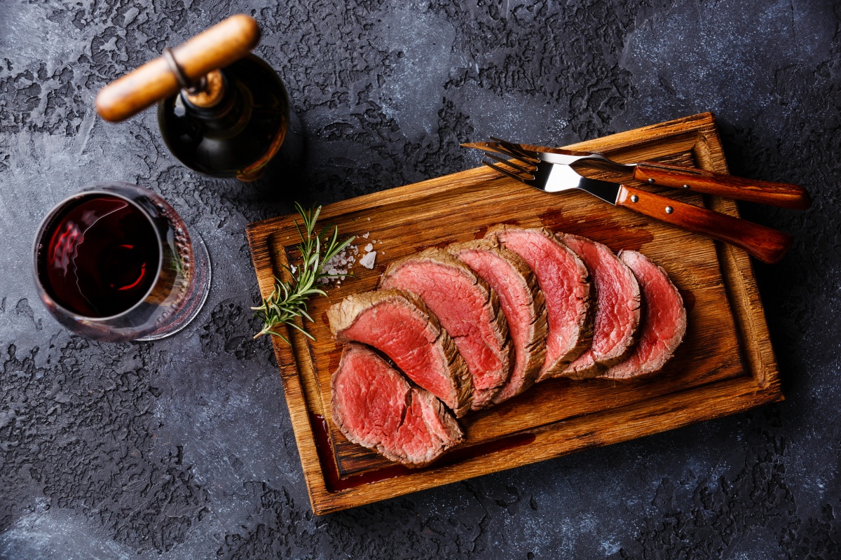 Medium-rare Rindersteak in Scheiben auf Holzbrett, Glas Rotwein und Besteck daneben, dunkle Tischoberfläche.