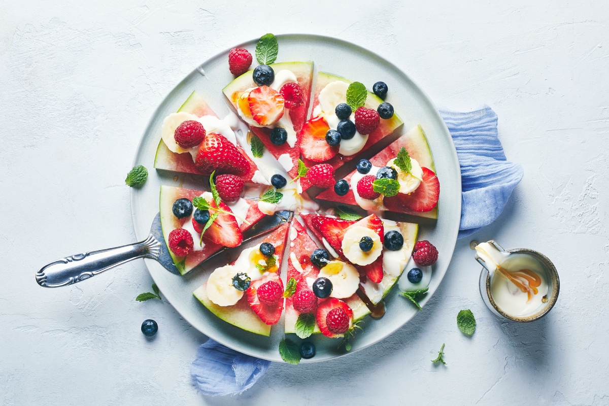 Eine fruchtige Melonen-Pizza liegt servierbereit auf einem Teller, daneben steht ein Krug mit Joghurt