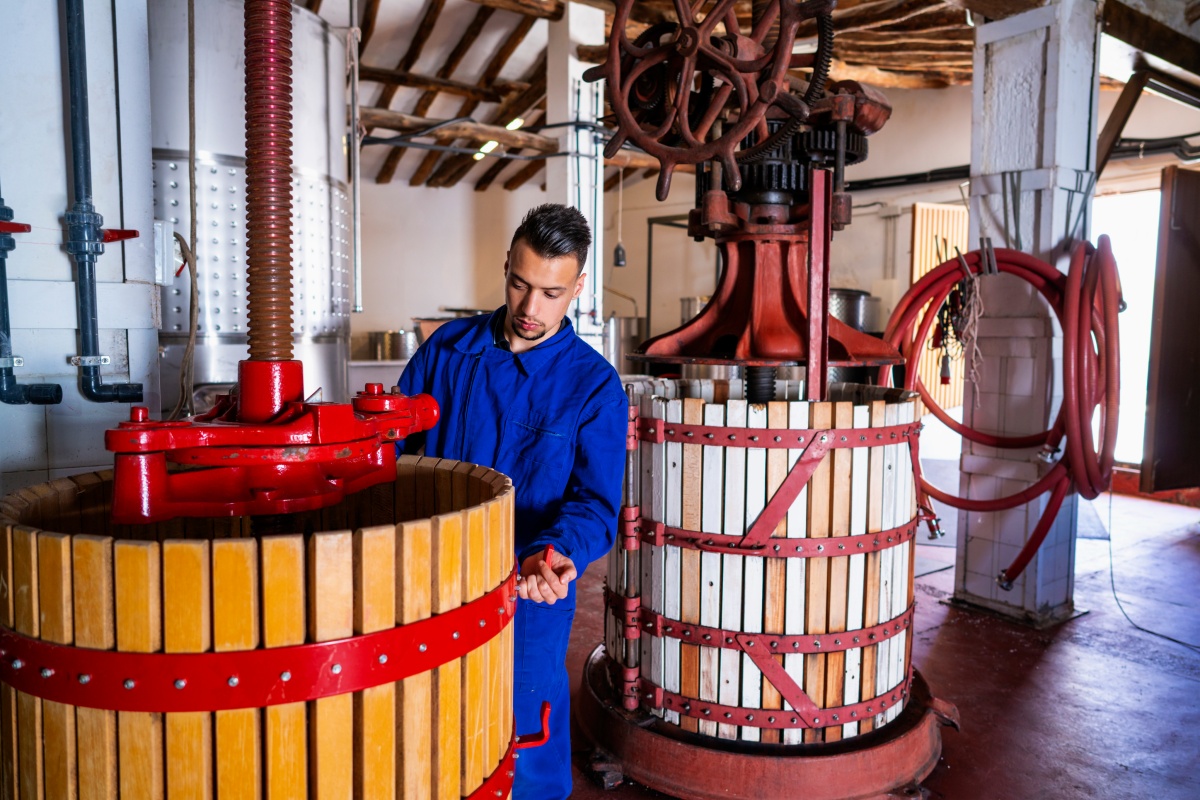 Mann in blauer Arbeitskleidung bedient traditionelle Weinpresse in einer Weinkellerei.
