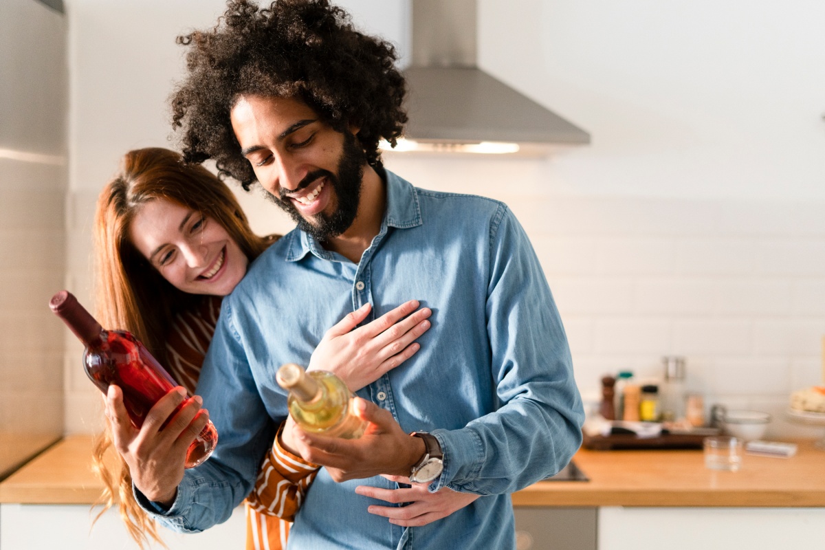 Lächelndes Paar in der Küche wählt zwischen zwei Weinflaschen die passende aus.
