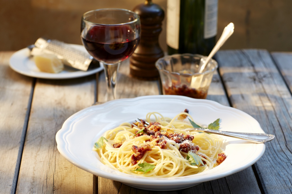 Holztisch mit Spaghetti-Teller und gefülltem Rotweinglas, dahinter unscharf Rotweinflasche, Gewürzmühle, Glasschale mit rotem Gewürz.