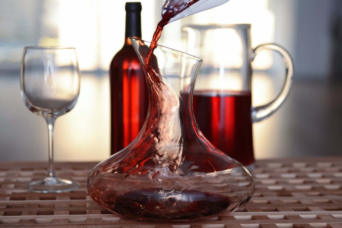 Rotwein wird in eine breite Glaskaraffe dekantiert. Im Hintergrund stehen eine Weinflasche, ein Weinglas und ein Krug.