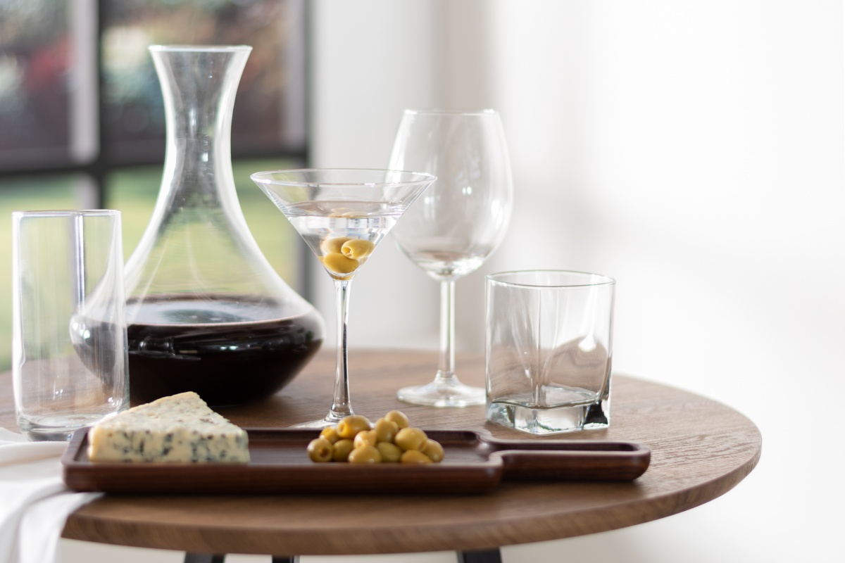 Eine stilvolle Anordnung einer mit Rotwein gefüllten Glaskaraffe, einem Martini-Glas mit Oliven, leeren Gläsern sowie Käse und Oliven auf einem Holztablett.