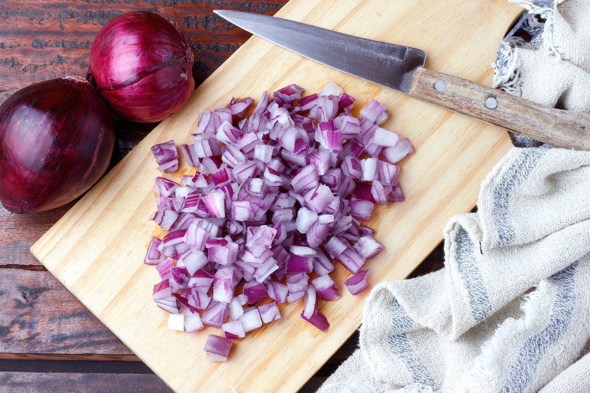 Gewürfelte rote Zwiebel auf einem Holzbrett neben einem Messer und zwei ganzen roten Zwiebeln.