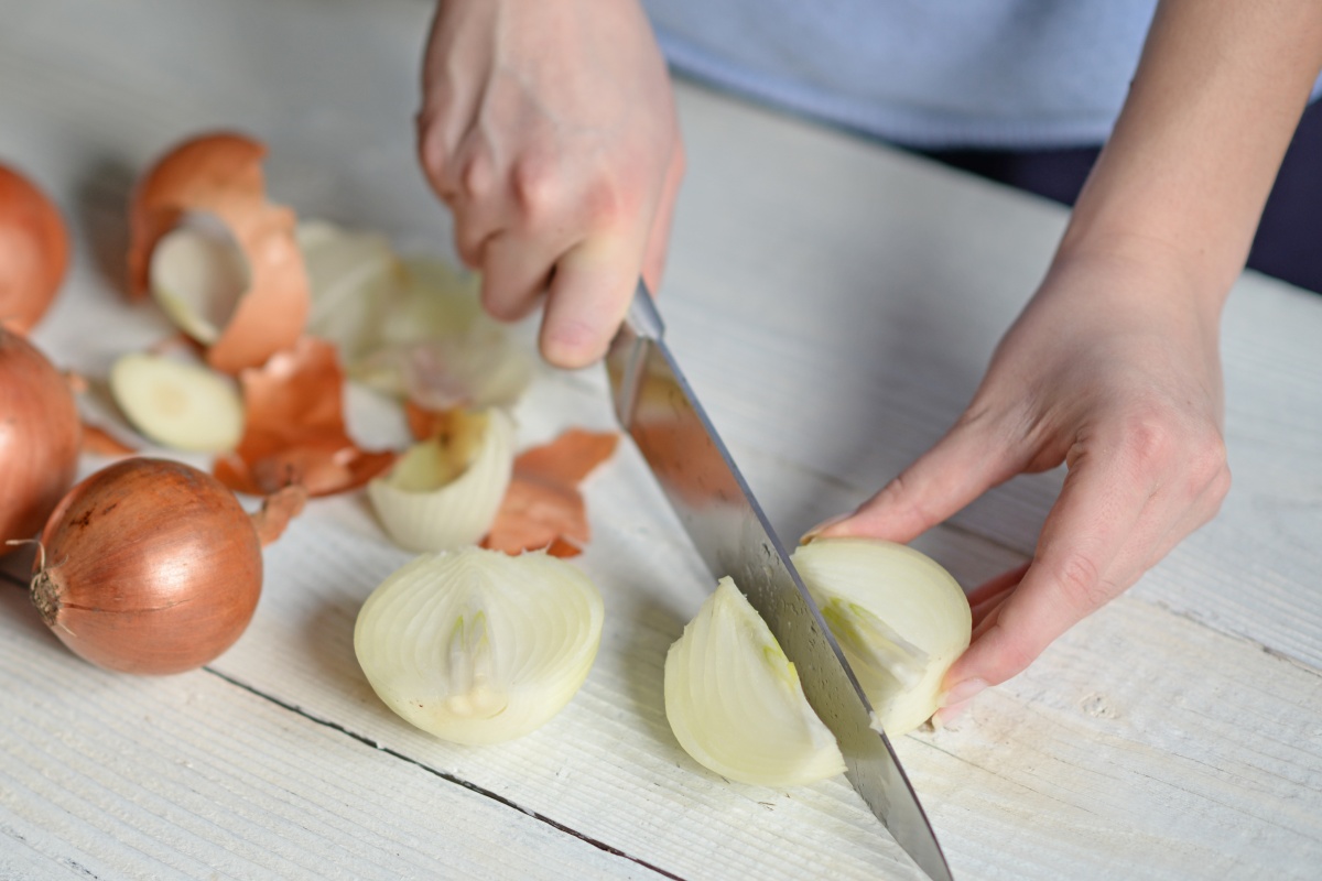 Person schneidet eine geschälte Zwiebel in Viertel auf einem weißen Tisch, daneben liegen Zwiebelschalen.