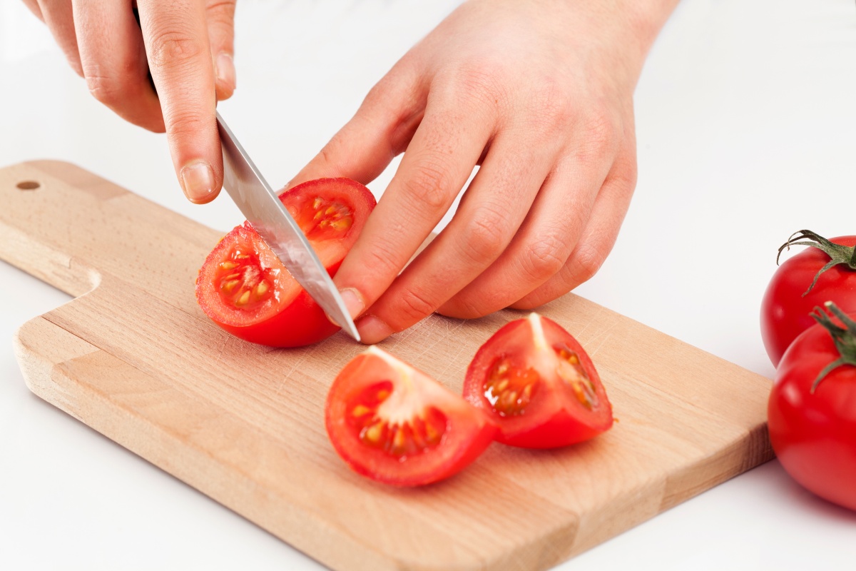 Person schneidet eine Tomate in Viertel auf einem Holzbrett, daneben liegen weitere ganze Tomaten.