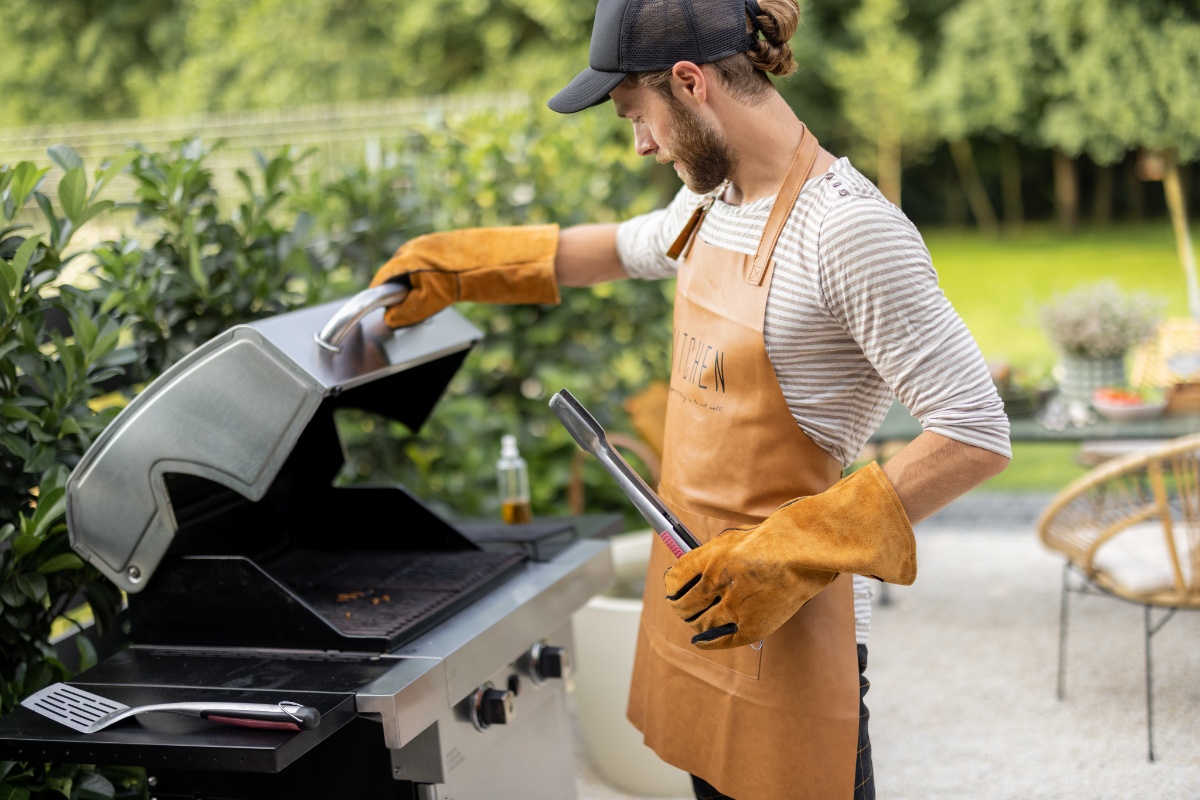Mann mit Grillhandschuhen, Schürze, Baseballcap und Grillzange in der Hand steht auf der Terrasse am Grill, dahinter Heckensträucher, Gartentisch und -stuhl, Wiese und Bäume.
