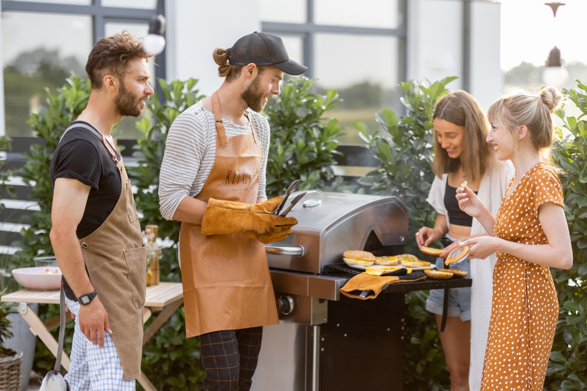 Zwei Männer in Grillschürze und zwei Frauen stehen um einen Gasgrill und bereiten Hamburger vor. Im Hintergrund kleiner Gartentisch aus Holz, Heckensträucher und dahinter ein Haus mit Glasfront.