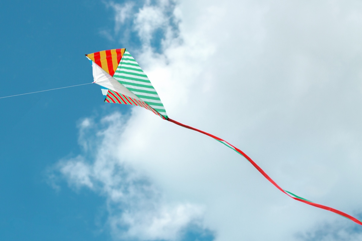 Selbst gebastelter Einleiner-Drachen mit roten und grünen Streifen fliegt hoch am blauen Himmel mit langen roten Bändern.