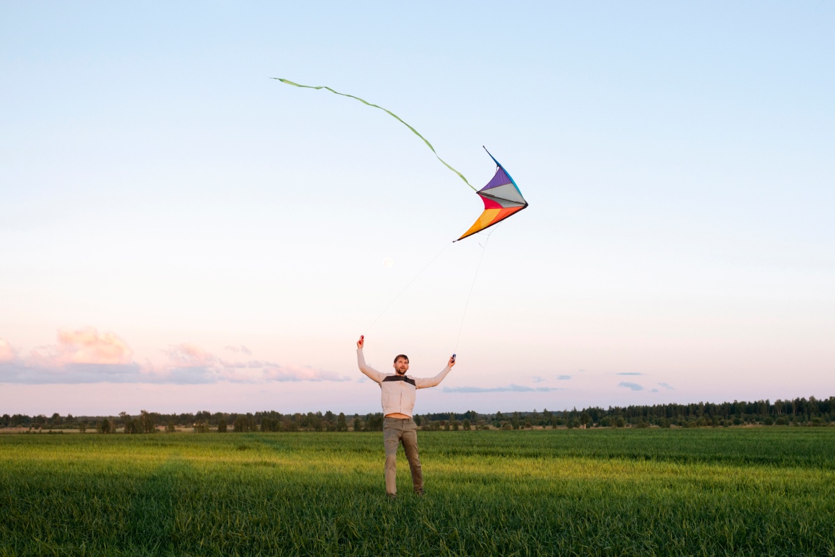 Mann fliegt einen bunten Lenkdrachen auf einer großen Wiese bei Sonnenuntergang, der Himmel ist leicht bewölkt.