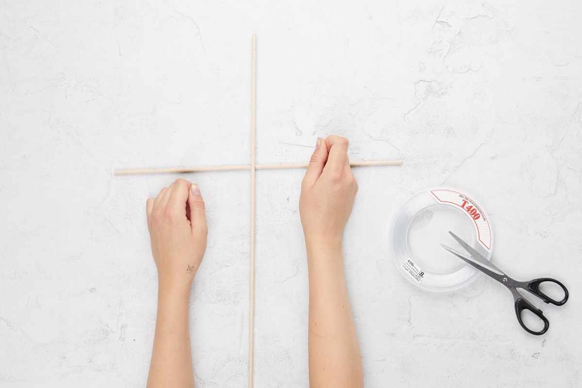 Hände binden zwei Holzstäbe zu einem Kreuz, um das Grundgerüst für einen Drachen zu basteln. Klebeband und Schere liegen bereit.