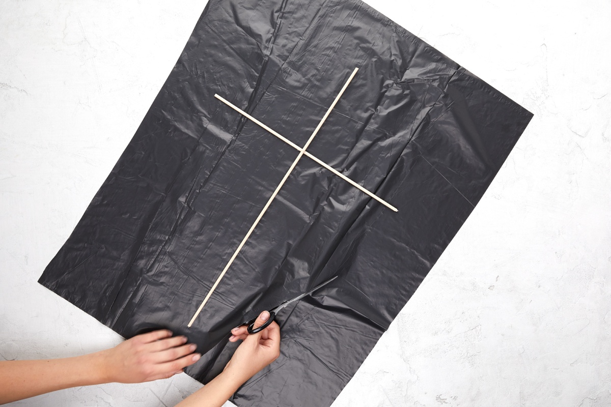 Holzkreuz auf schwarzer Mülltüte, Hände schneiden Mülltüte in Form eines Drachens aus.