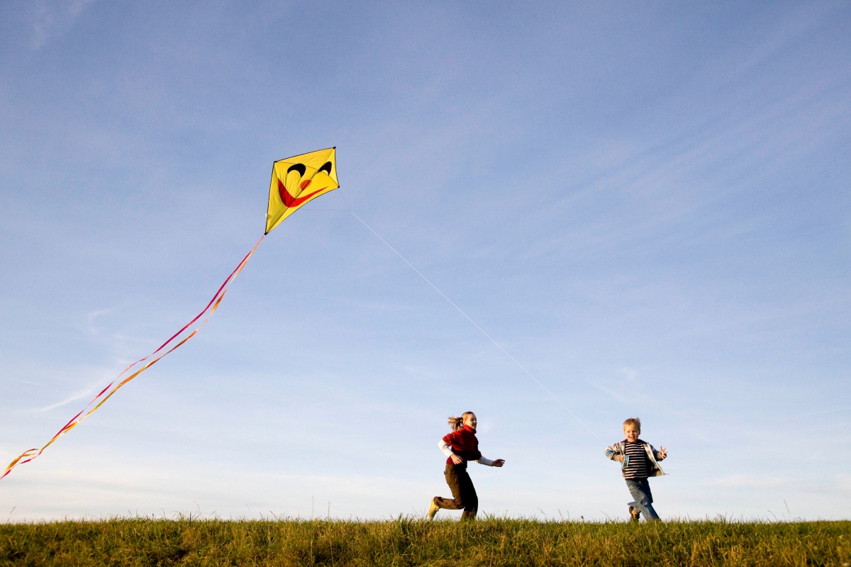 Zwei Kinder laufen auf einer Wiese und lassen einen gelben Drachen mit fröhlichem Gesichtsmotiv und bunten Bändern fliegen.