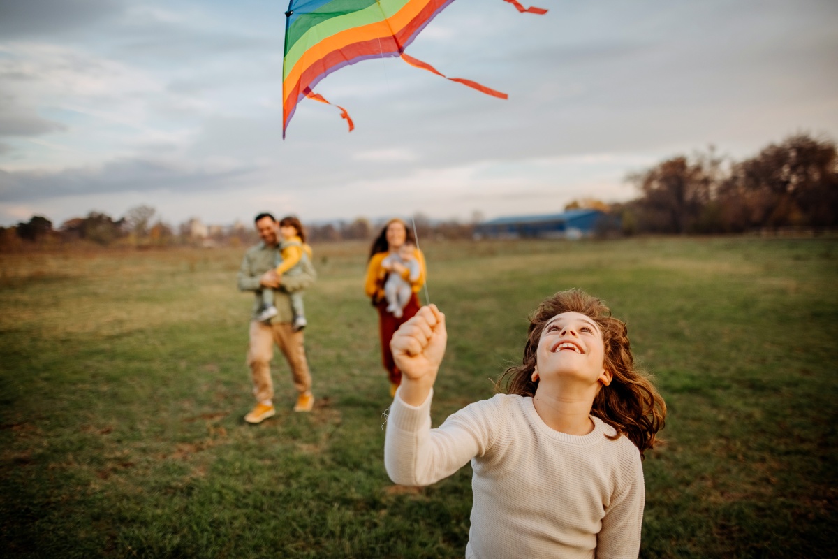 Kind lässt einen bunten Drachen auf einer Wiese fliegen, während die Familie im Hintergrund fröhlich zuschaut.