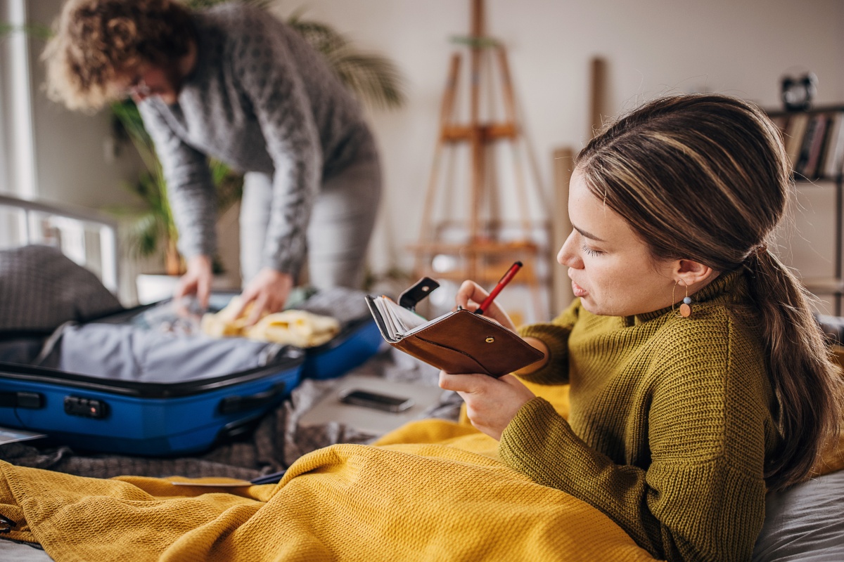 Frau schreibt eine Packliste in ein Notizbuch, während ein Mann Kleidung in einen Koffer packt.