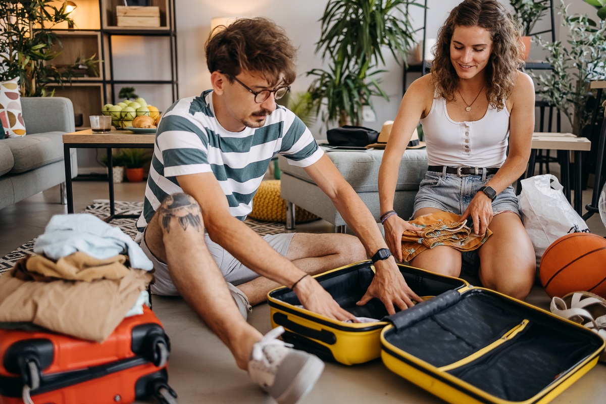 Paar sitzt auf dem Boden und packt einen gelben Koffer, umgeben von Kleidung, Schue und anderen Gegenständen.