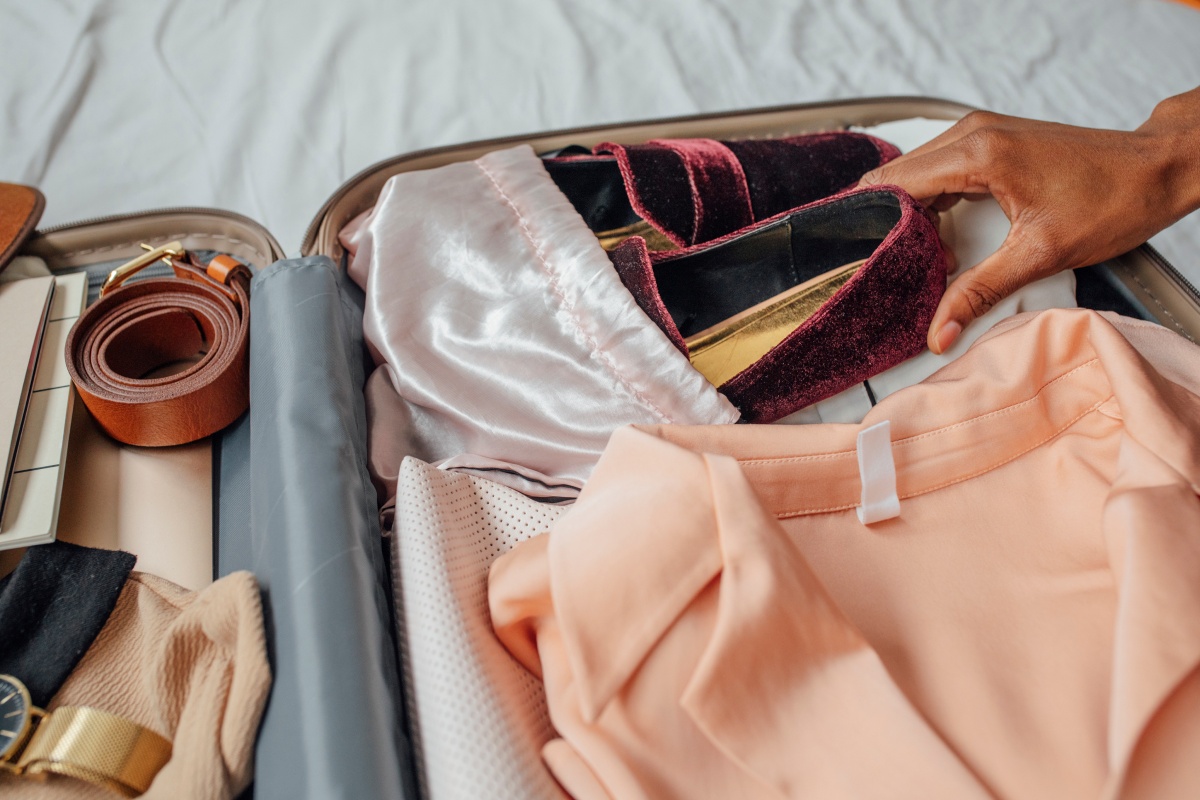 Person packt elegante, weinrote Schuhe in einen Koffer mit Kleidung, Gürtel und Accessoires.