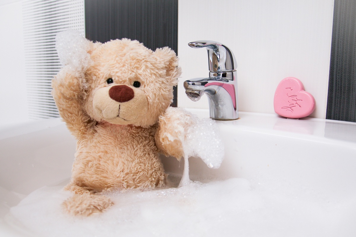 Ein Teddybär sitzt im Schaum in einem Waschbecken und wird von Hand gewaschen.
