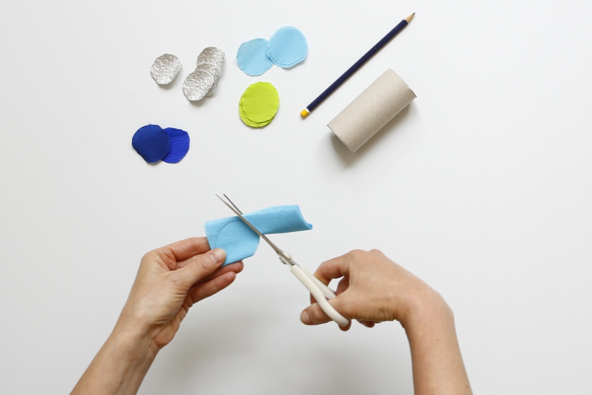 Jemand schneidet mit den Händen blaue Krepppapierschuppen, daneben bunten Schuppen, Stift und Papprolle auf weißem Hintergrund.