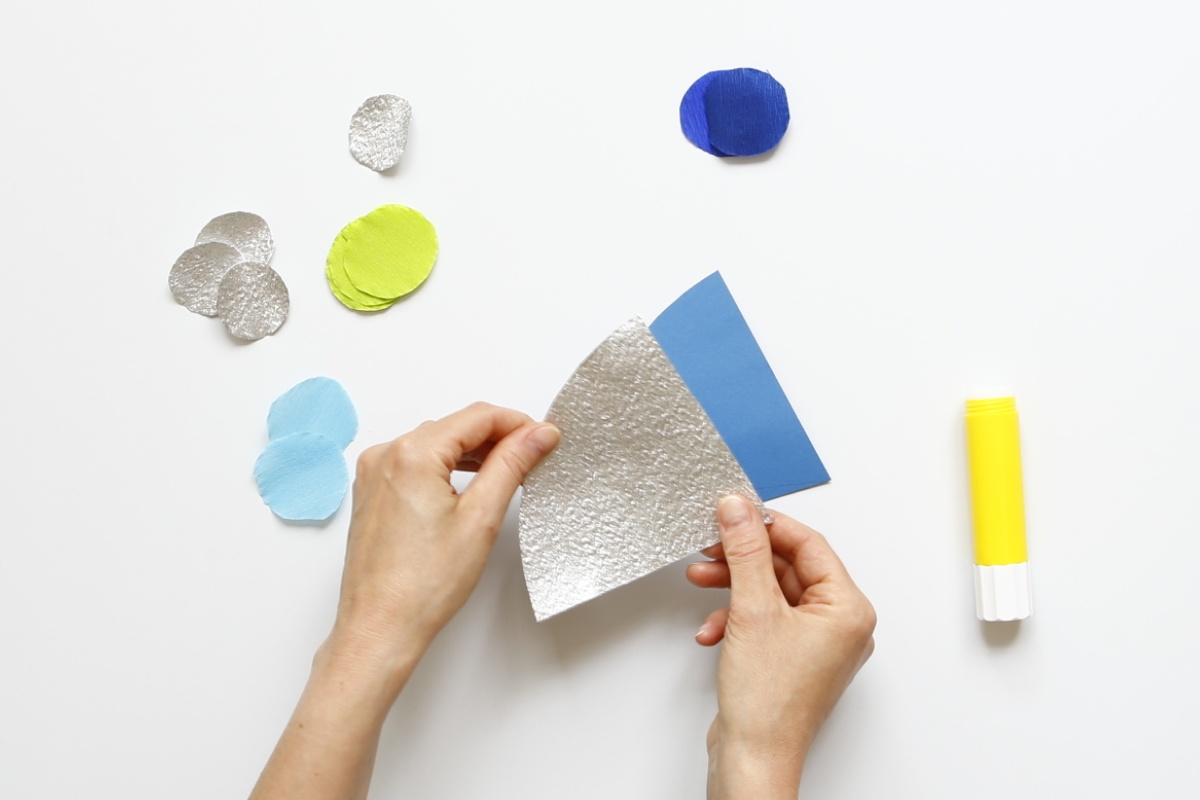 Hände halten blaue und silberne Papierschuppen, daneben farbige Schuppen und gelber Klebestift.