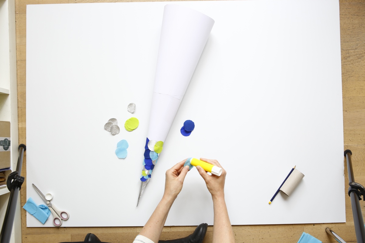 Hände kleben bunte Papierschuppen auf weiße Schultüte.