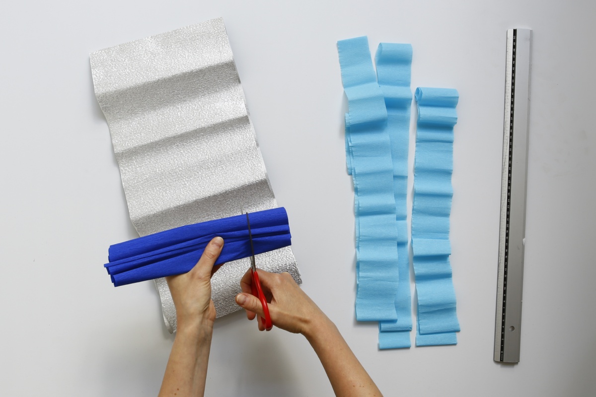 Hände schneiden blaues Krepppapier, neben silbernem Papier und blauen Papierstreifen.
