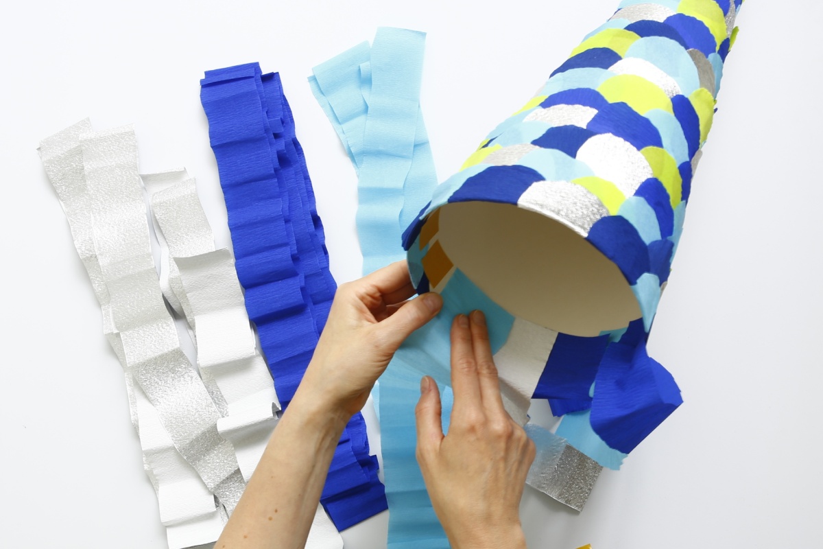 Hände kleben blaue und silberne Krepppapierstreifen auf eine Schultüte mit bunten Schuppen.