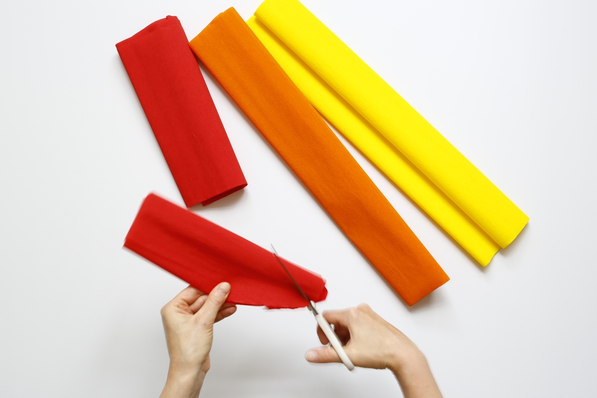 Hände schneiden rotes Krepppapier, daneben liegen orange und gelbe Krepppapierstreifen.