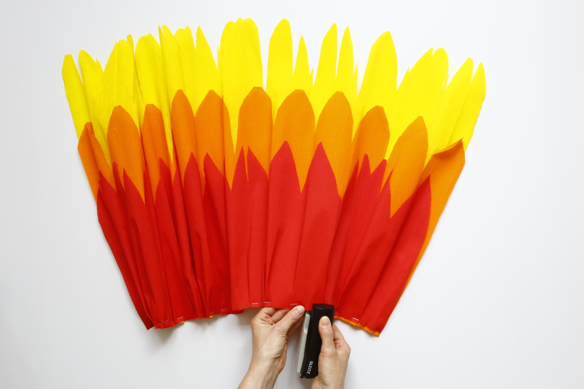 Hände tackern gelbe, orange und rote Krepppapierstreifen zusammen, die wie Flammen aussehen.