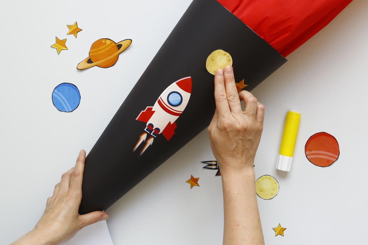 Hände kleben Weltraum-Motive wie Rakete und Planeten auf schwarze Schultüte mit rotem Krepppapier.