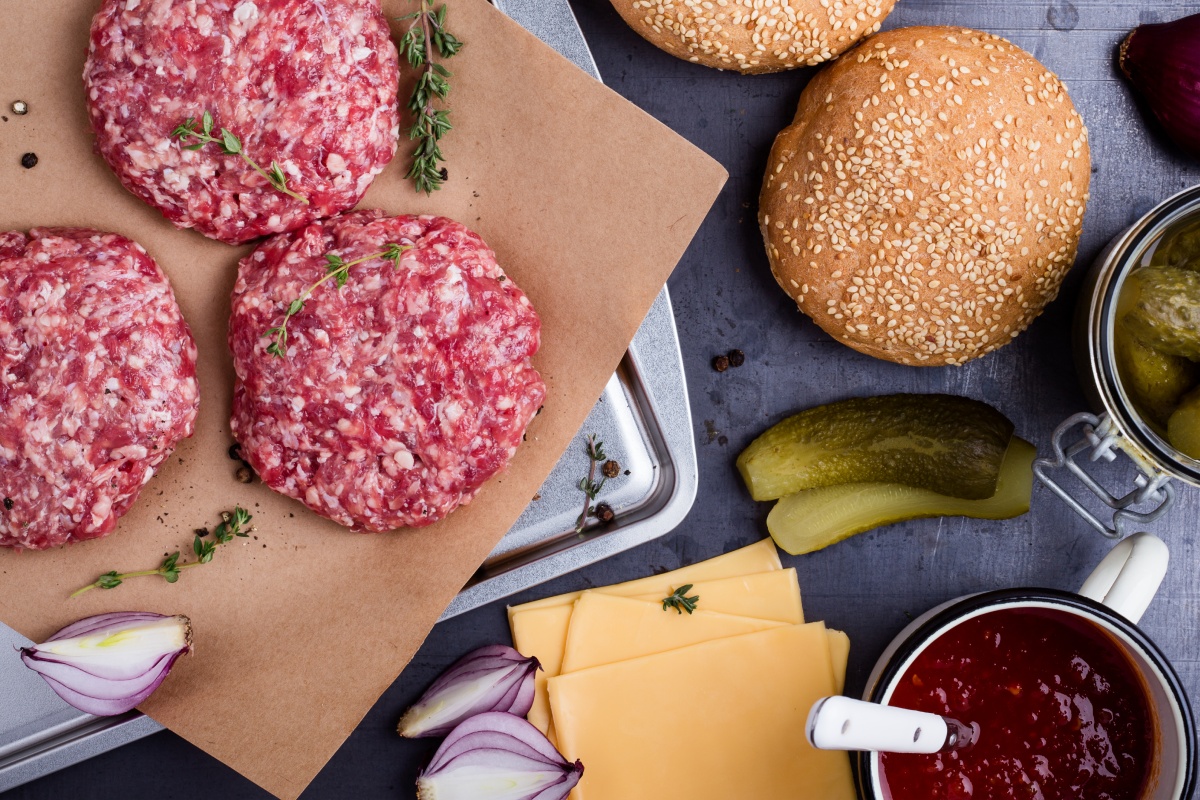 Burger-Zutaten wie rohe Patties, Sesambrötchen, Käse, Gurken, Zwiebeln und Ketchup liegen auf einem Tisch.