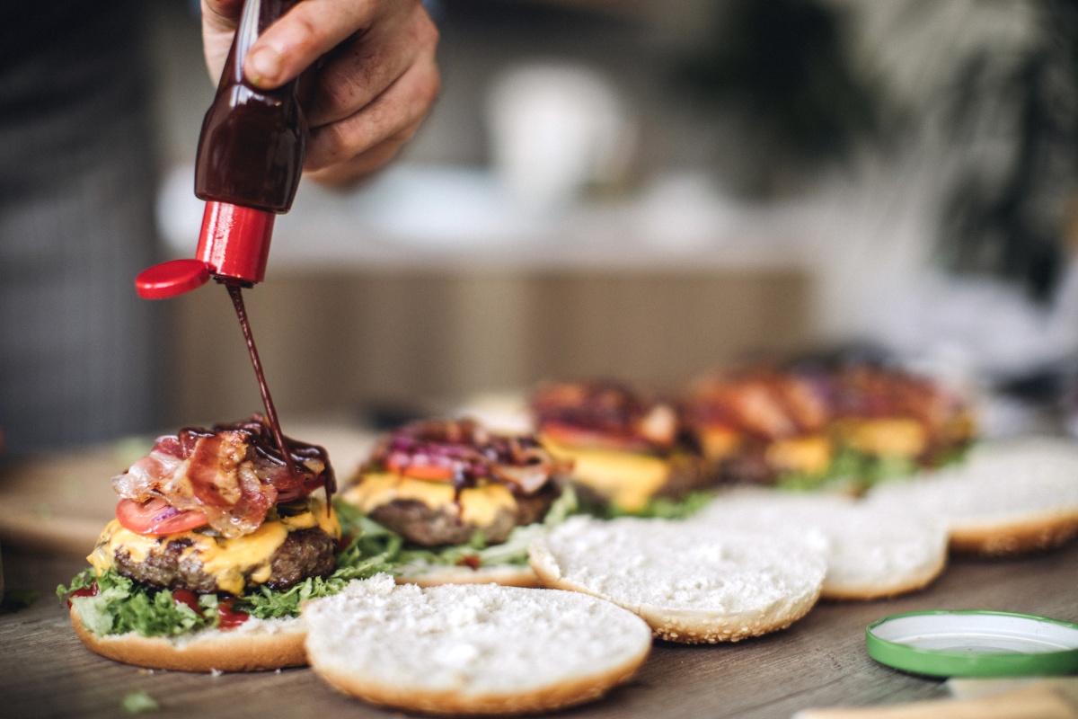 Eine Person würzt vier geöffnet daliegende Burger mit Barbecue-Sauce.