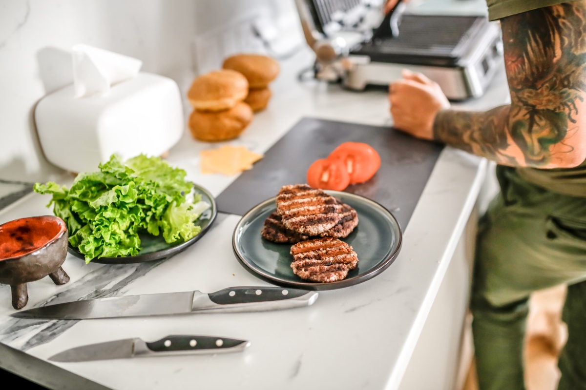 Gegrillte Burger-Patties auf einem Teller, daneben liegen Salat, Tomaten, Brötchen und Messer auf einer Küchenarbeitsplatte.