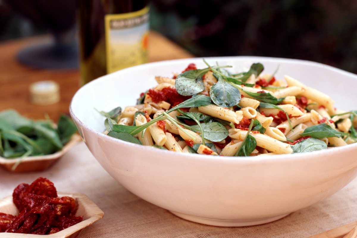 Nudelsalat mit getrockneten Tomaten und frischem Blattspinat in einer weißen Keramikschüsse.