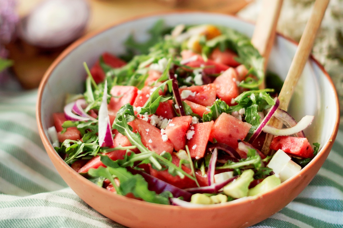 Schüssel mit frischem Salat aus Wassermelone, Rucola, roten Zwiebeln und Feta auf gestreifter Tischdecke.