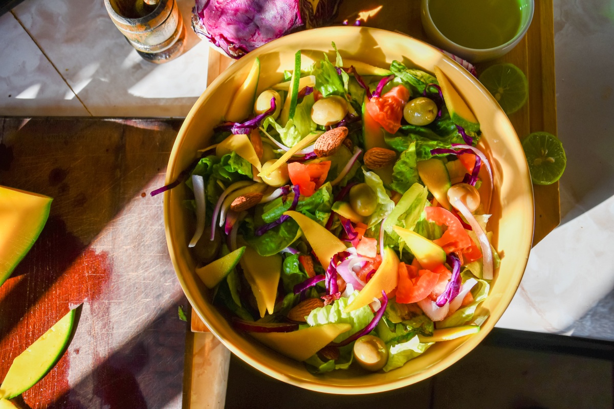 Schüssel mit gemischtem Salat aus Tomaten, Mandeln, Mango, Oliven, roten Zwiebeln und grünem Salat auf Holztisch.