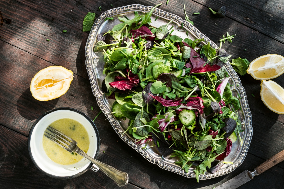 Frischer gemischter Salat auf einem silbernen Tablett, daneben Zitronenhälften und eine Schüssel mit Dressing auf Holztisch.