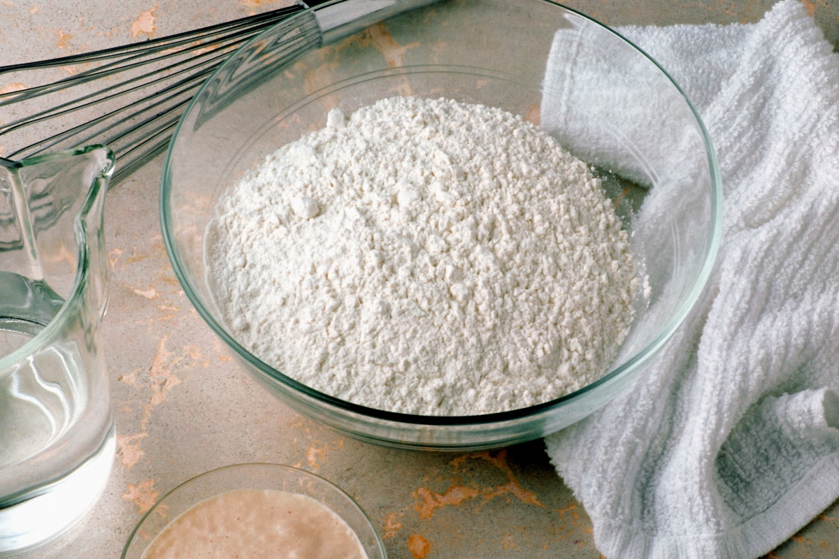 Eine Glasschüssel mit Mehl, daneben ein Behältnis mit Wasser und Sauerteig auf einer Küchenarbeitsfläche.